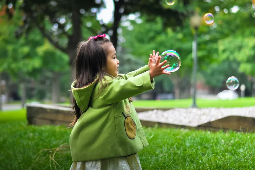 little girl in green coat playing with bubbles in park.
