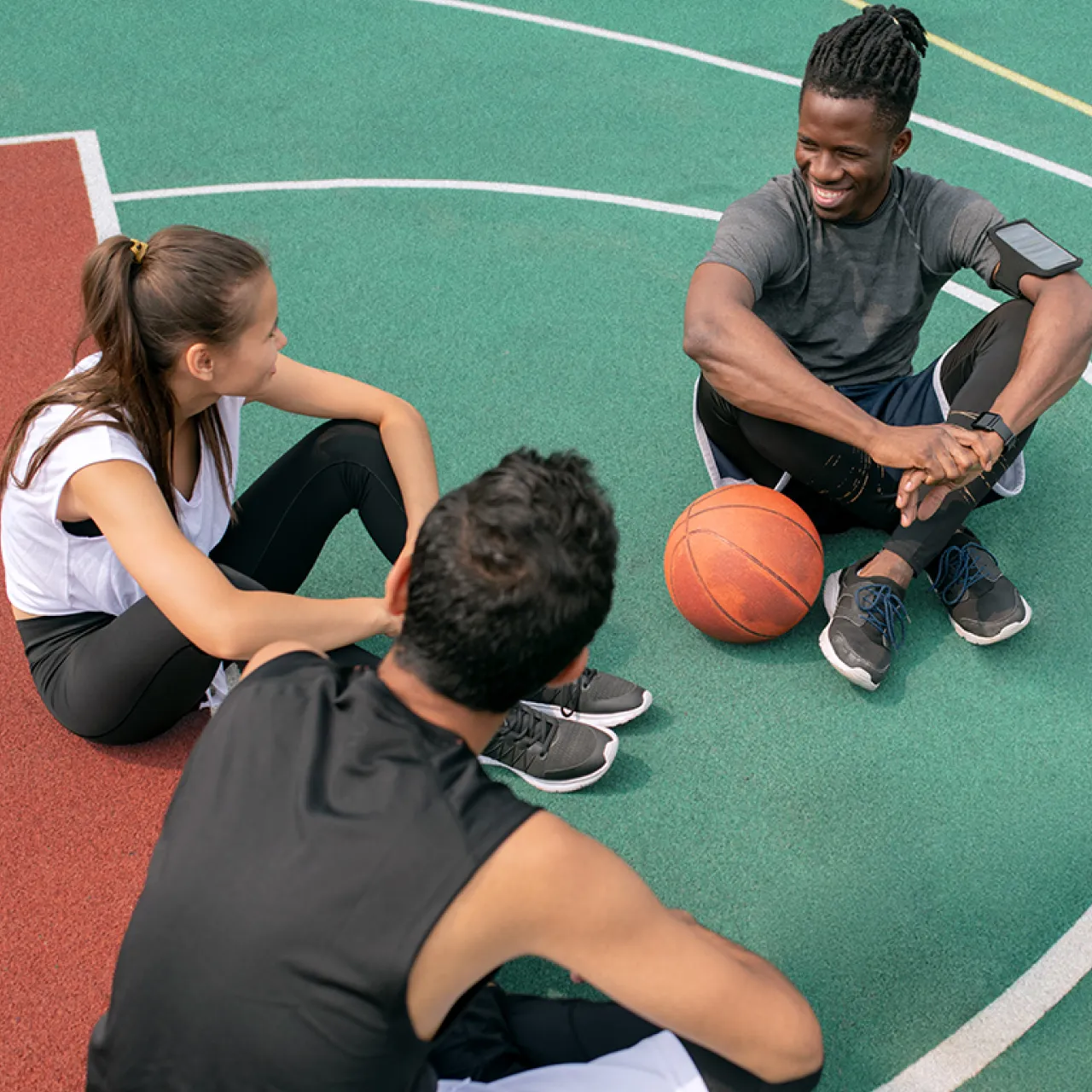 three-teens-sitting-on-rubber-based-basketball-court-smiling