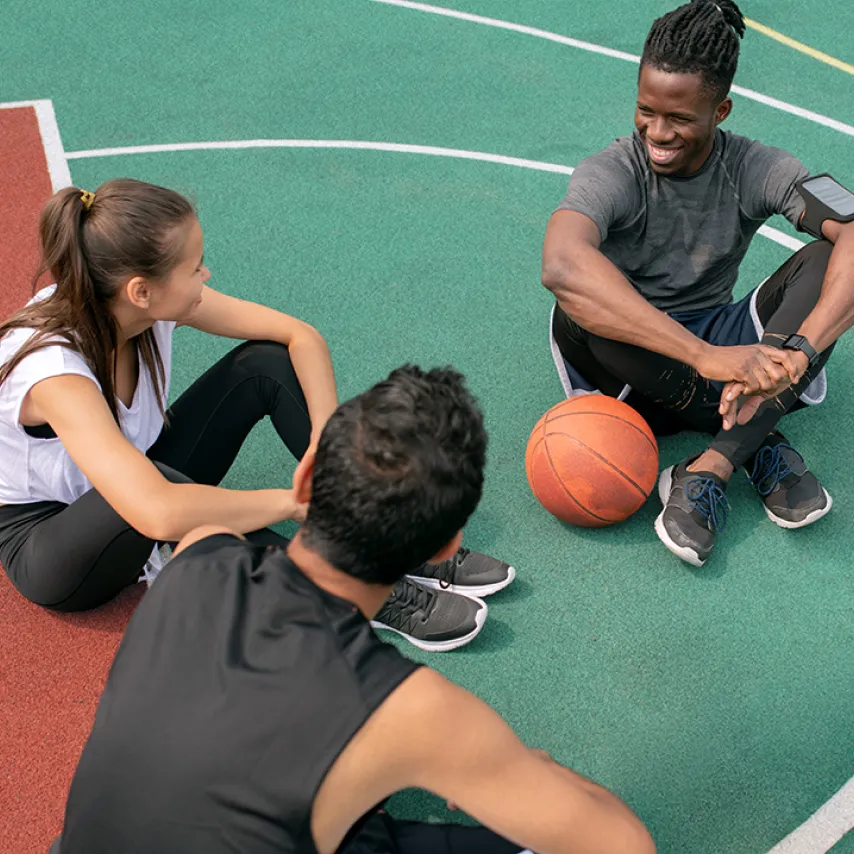 three teens sitting on rubber based basketball court smiling.
