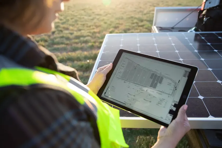 worker looking at solar farm plan on tablet next to uninstalled solar panel.
