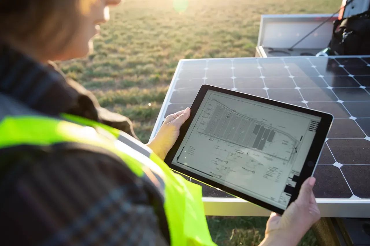 worker looking at solar farm plan on tablet next to uninstalled solar panel.