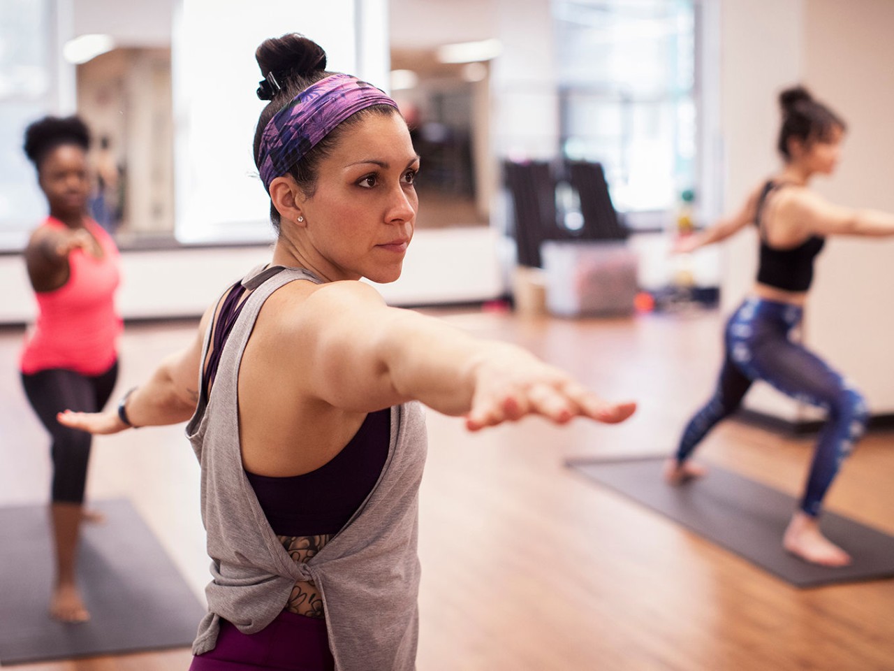 three women practice yoga in a brightly lit studio, holding a standing pose on their mats.