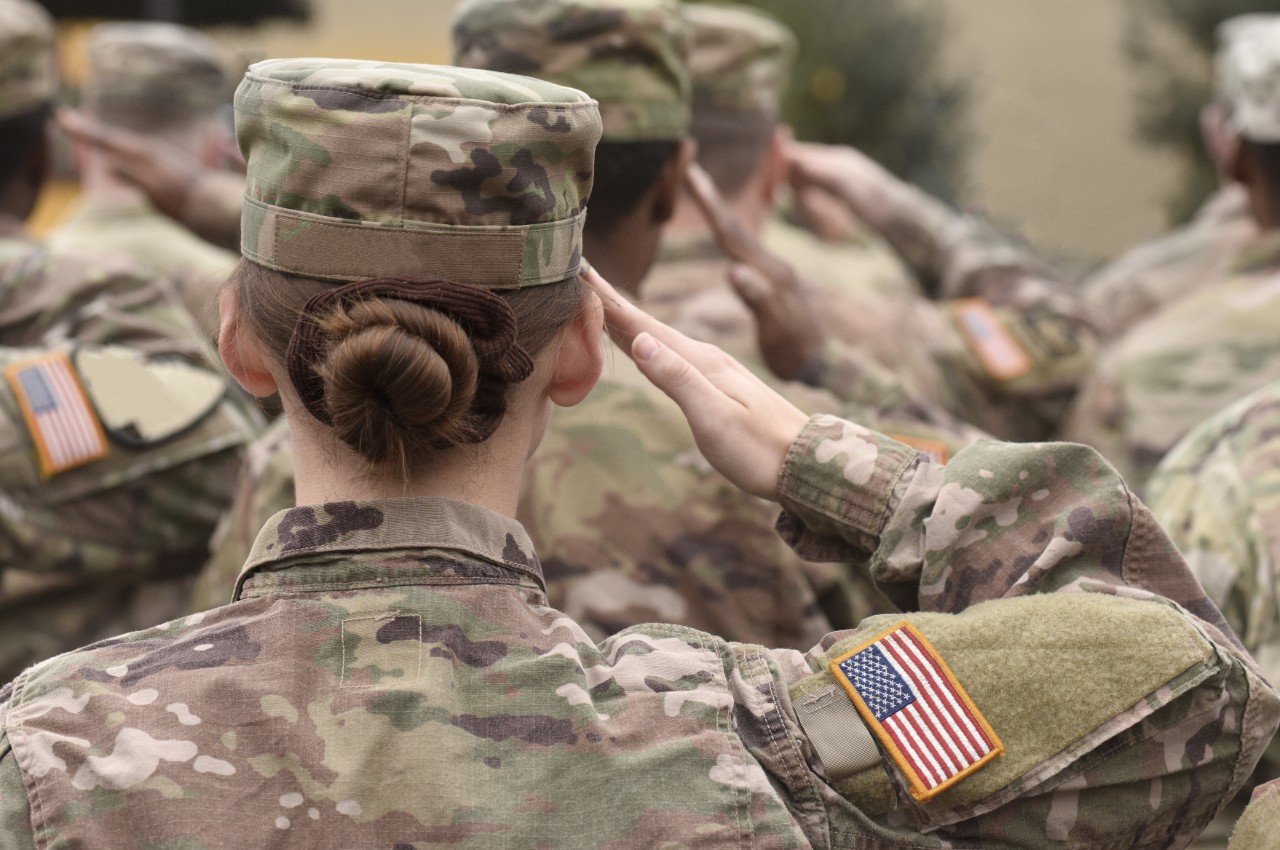 U.S. soldiers in uniform salute; focus on a woman with a bun and a flag patch on her sleeve.