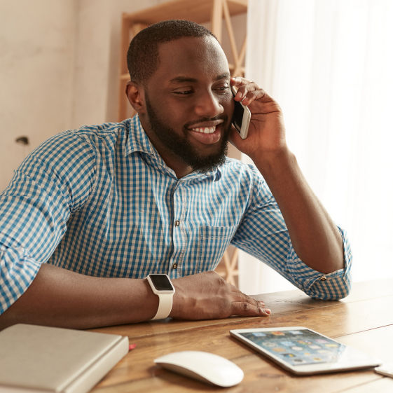 smiling man in a blue checked shirt talks on the phone at a desk with a tablet, mouse, and notebook.