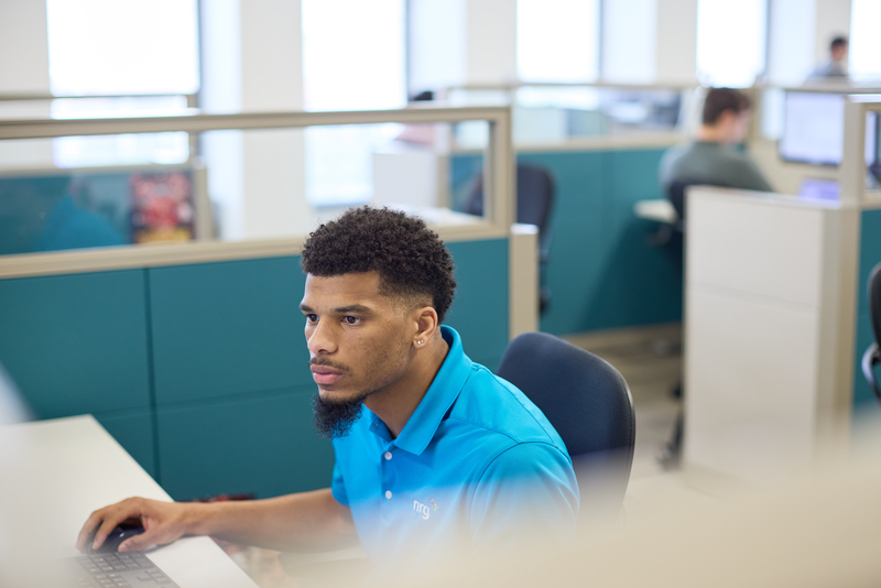 man in blue shirt working at a computer in a modern office with cubicles and colleagues in the background.