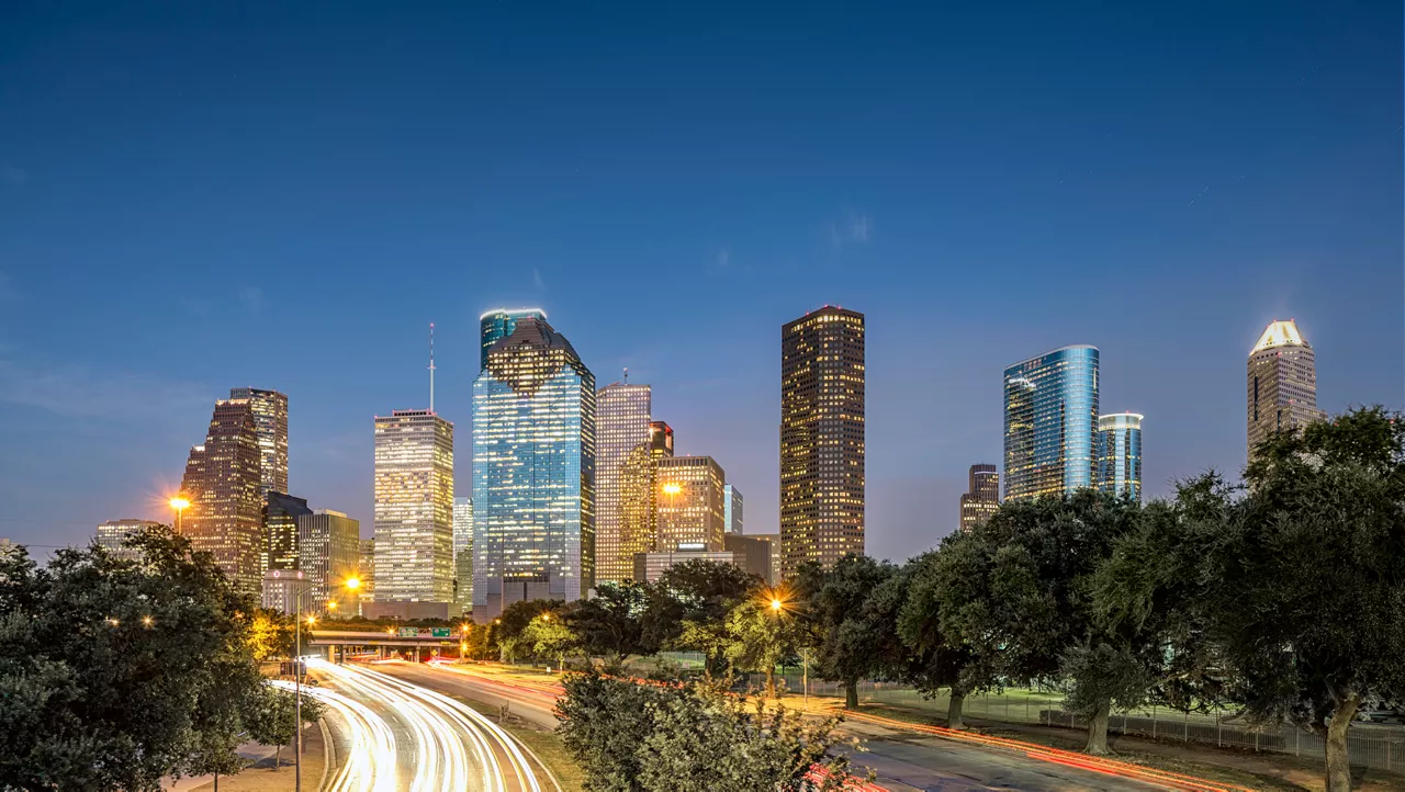 Houston skyline at dusk with tall buildings, trees, and light trails from traffic in the foreground.