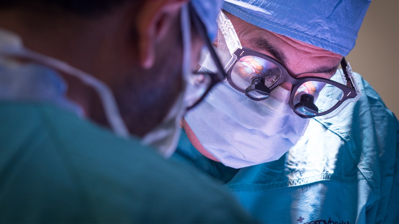 two surgeons in scrubs operate, one wears surgical loupes and a mask, focused under bright operating lights.