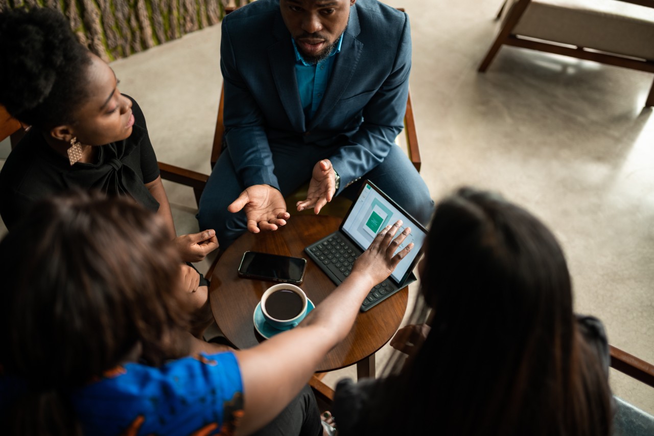 three colleagues meeting at a small table with a tablet, keyboard, phone, and coffee.
