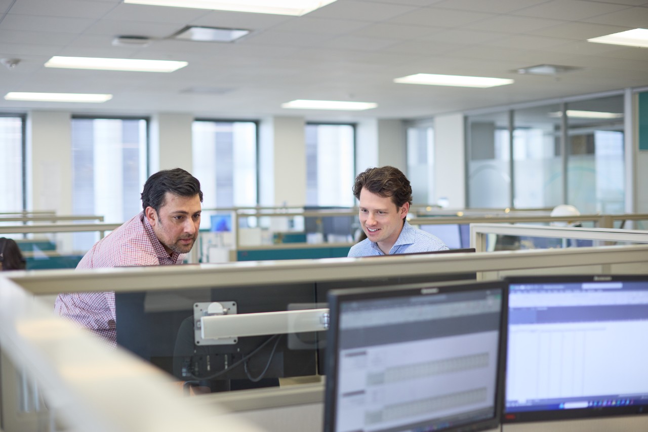 two men in an office look at a computer screen together in a row of cubicles with monitors.
