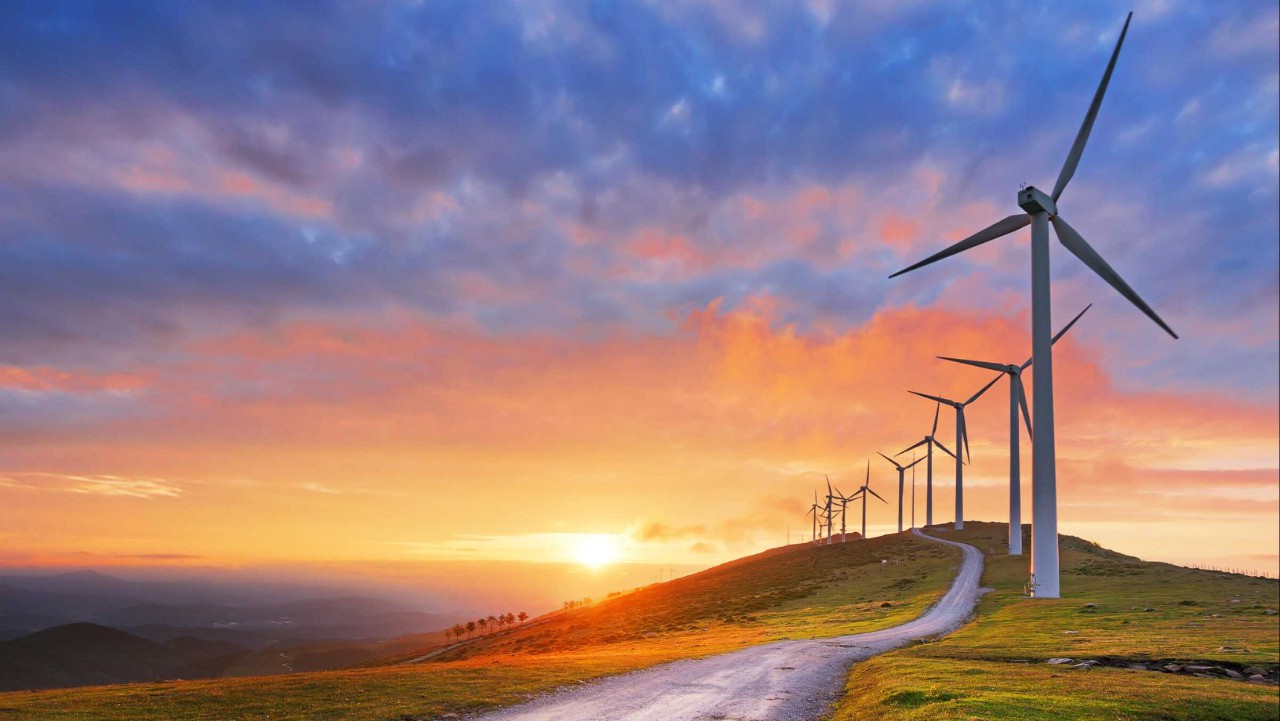 wind turbines on a hill at sunset, with a winding road leading toward the glowing horizon.