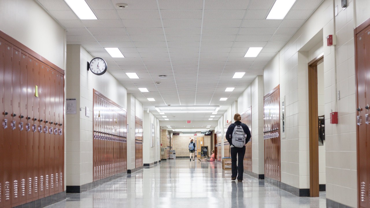 school hallway with brown lockers on both sides, a few students walking, and a clock on the wall.