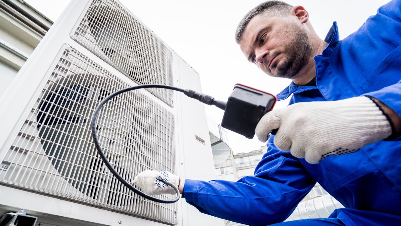 technician in blue coveralls inspects an outdoor HVAC unit with a camera tool.