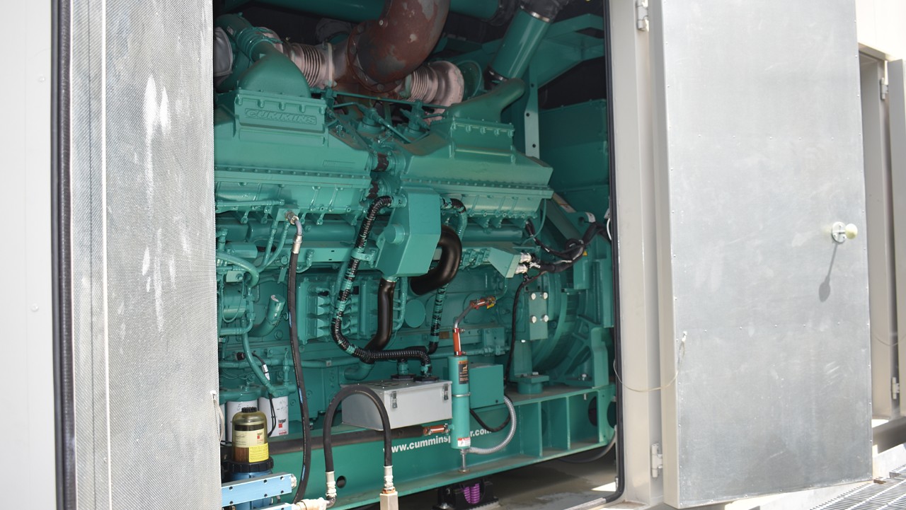 large green industrial engine inside a metal enclosure with open doors, showing pipes and wiring.
