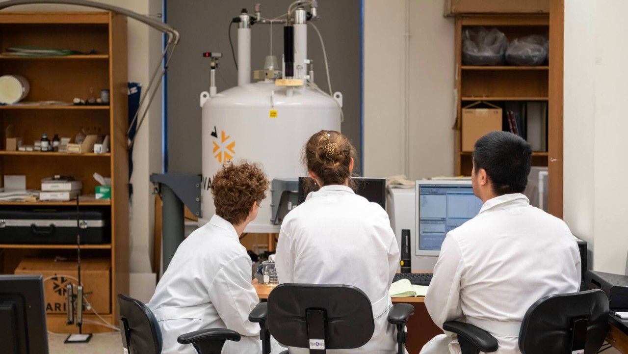 three people in lab coats work at computers facing scientific equipment in a laboratory.