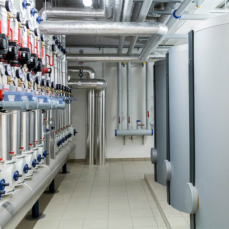 industrial boiler room with large pipes, gauges, and grey tanks on a tiled floor.