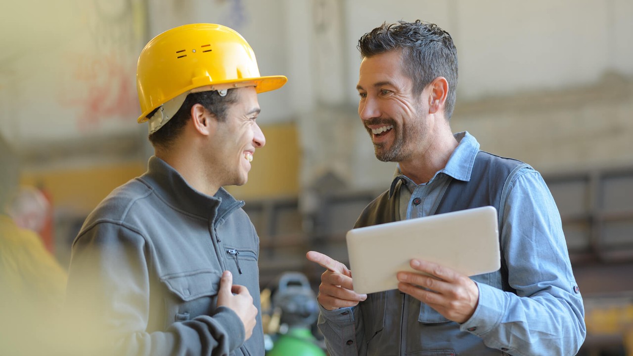 two men at a construction site, one wearing a hard hat, smiling and talking while holding a tablet.