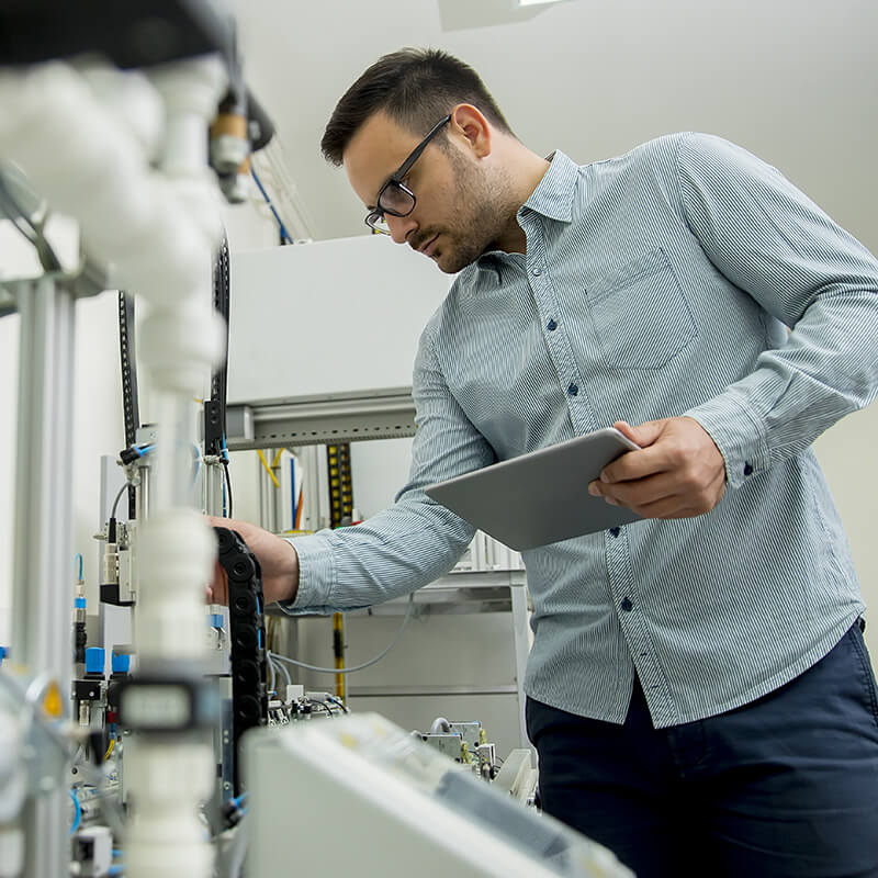 man in glasses holds a tablet while inspecting electronic equipment in a laboratory setting.