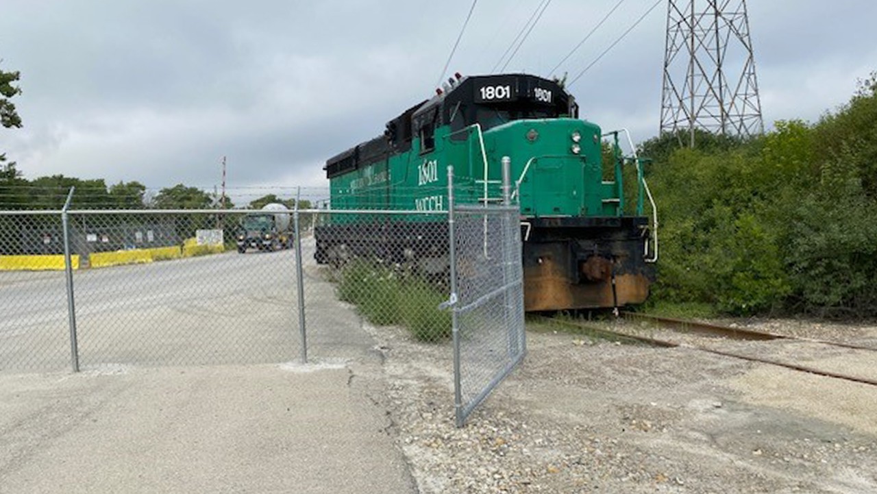 green locomotive on train tracks behind a chain-link fence, with trees and cloudy sky in the background.