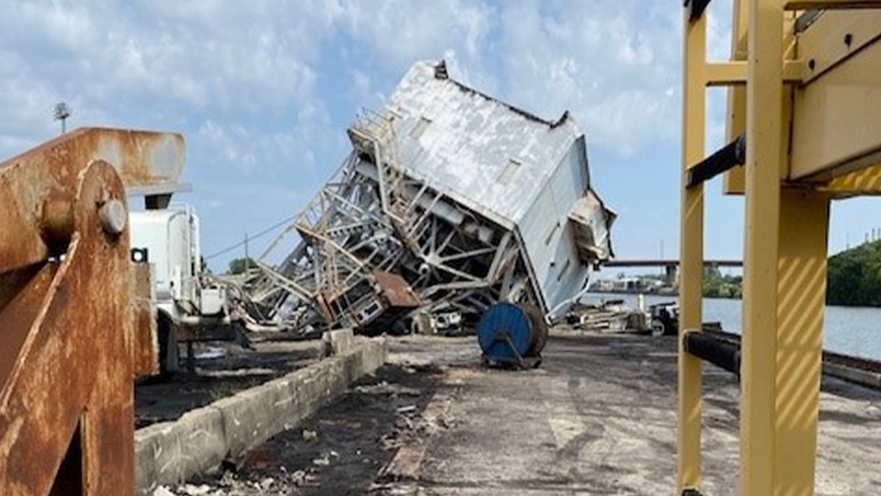 a large, damaged metal building is tilted and partially collapsed near water under a blue sky with clouds.