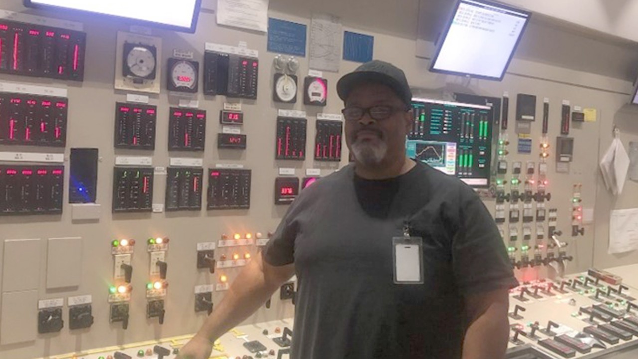 man in a control room stands by a control panel with illuminated buttons, screens, and gauges.