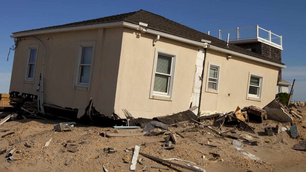 a house tilted and damaged, surrounded by debris, after a natural disaster.