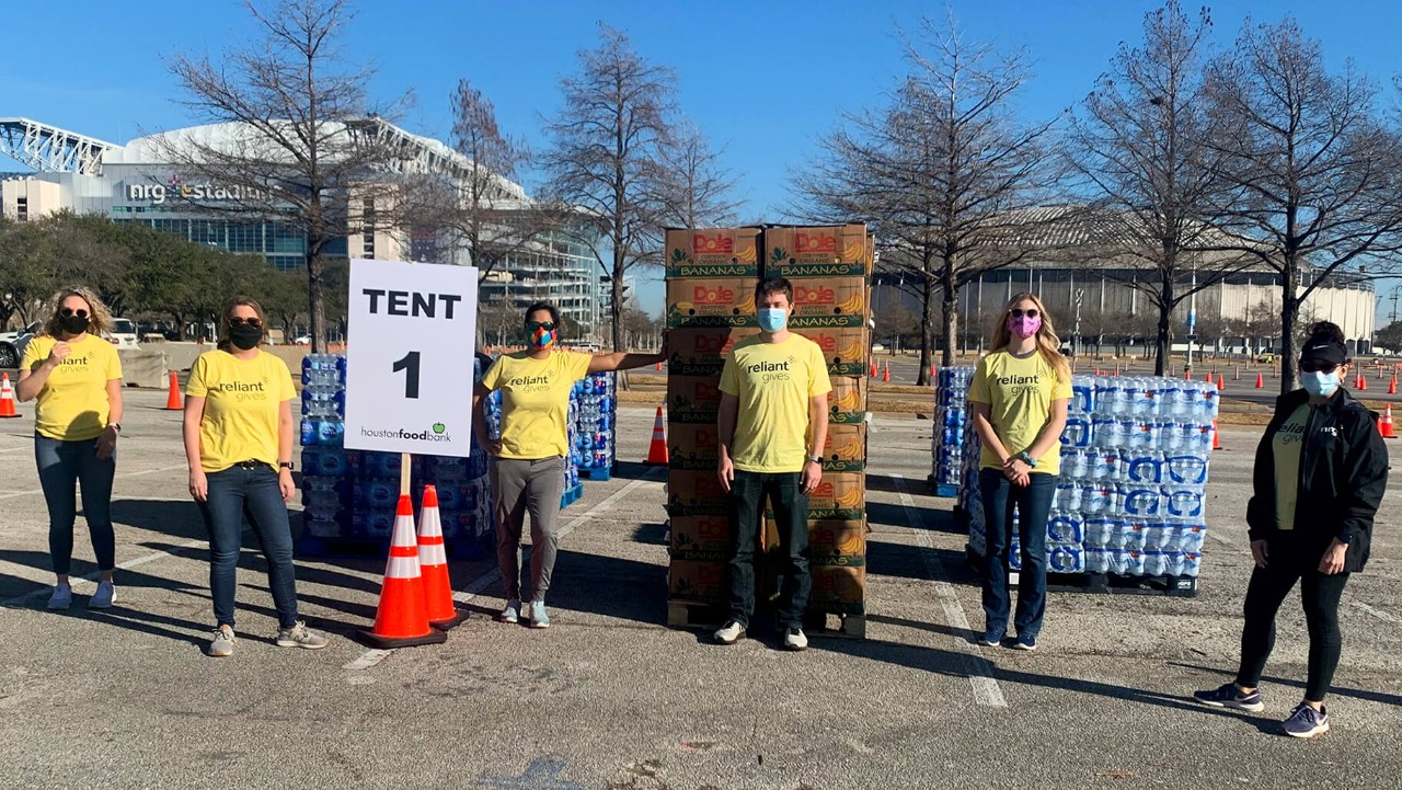 six people in yellow shirts stand by bottled water, food boxes, and a Tent 1 sign in a parking lot.