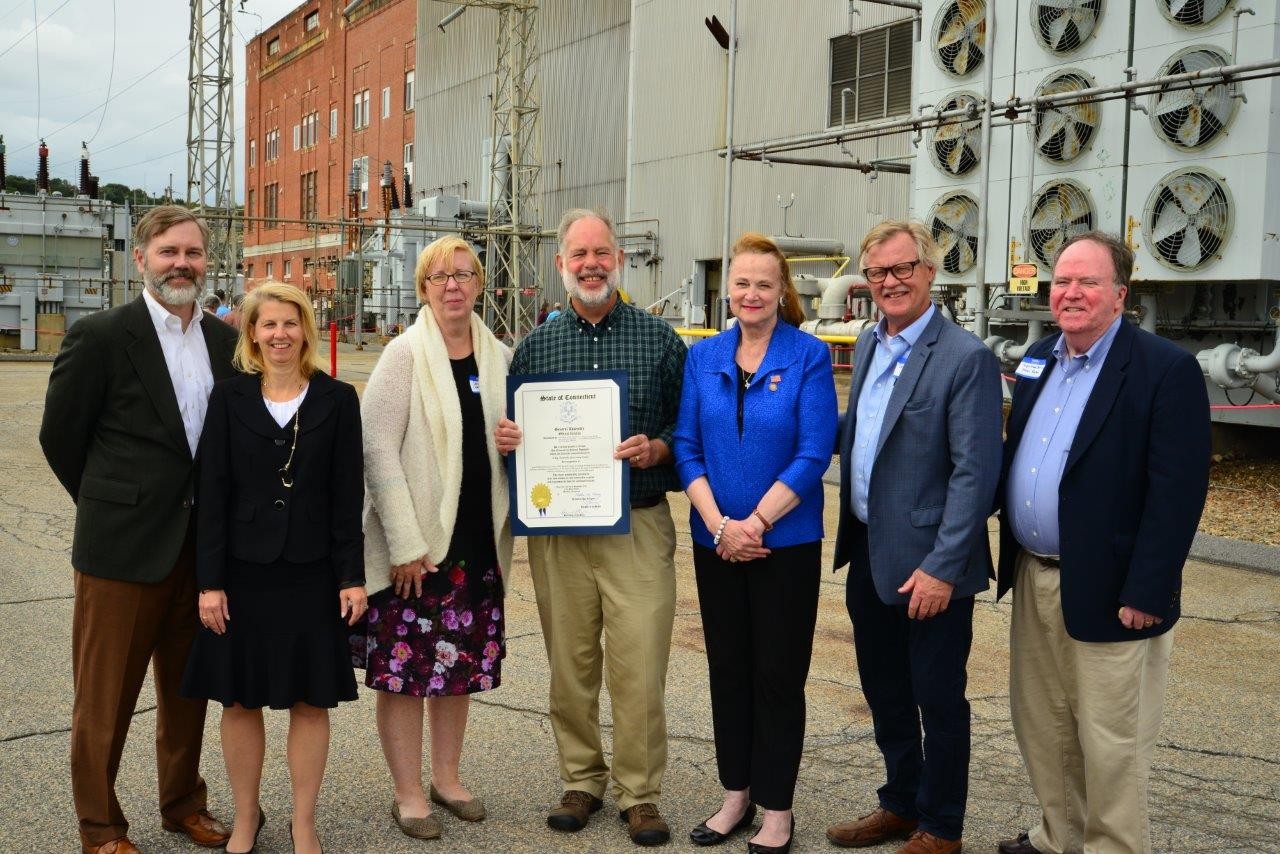 seven adults stand outdoors near industrial buildings, one holding an award certificate and smiling at the camera.