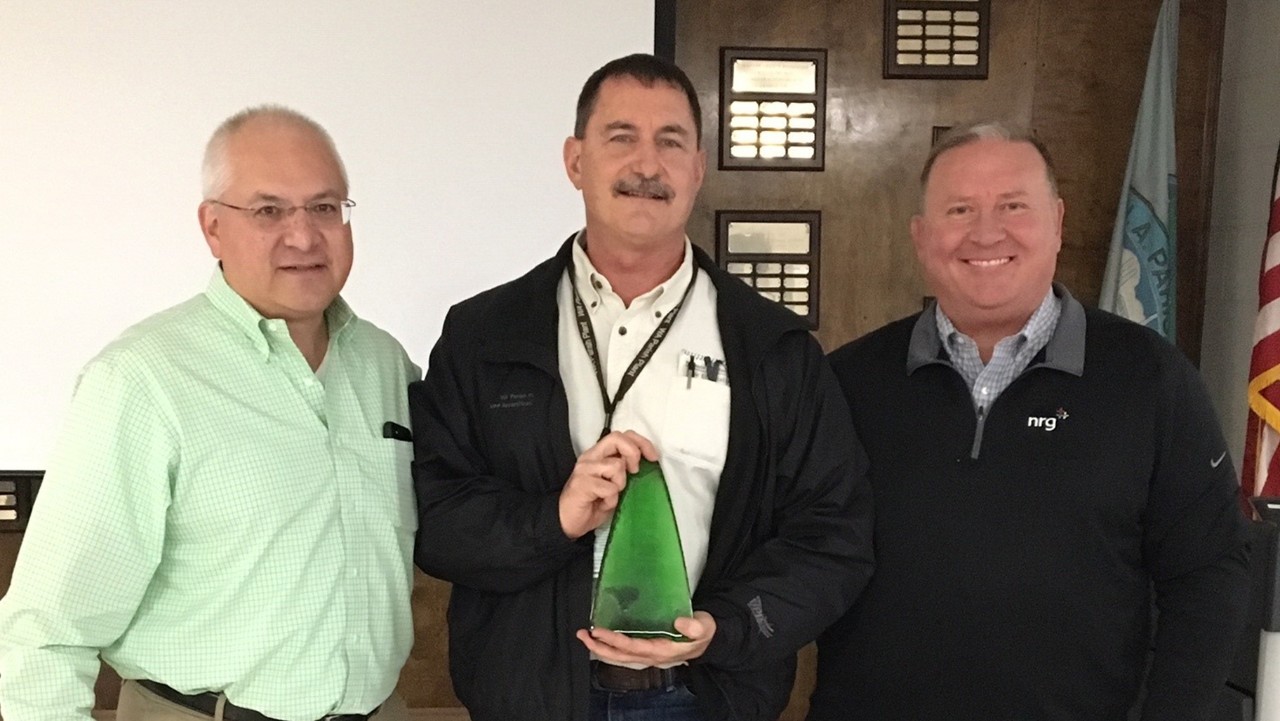 three men standing indoors, the center man holding a green award, all smiling at the camera.