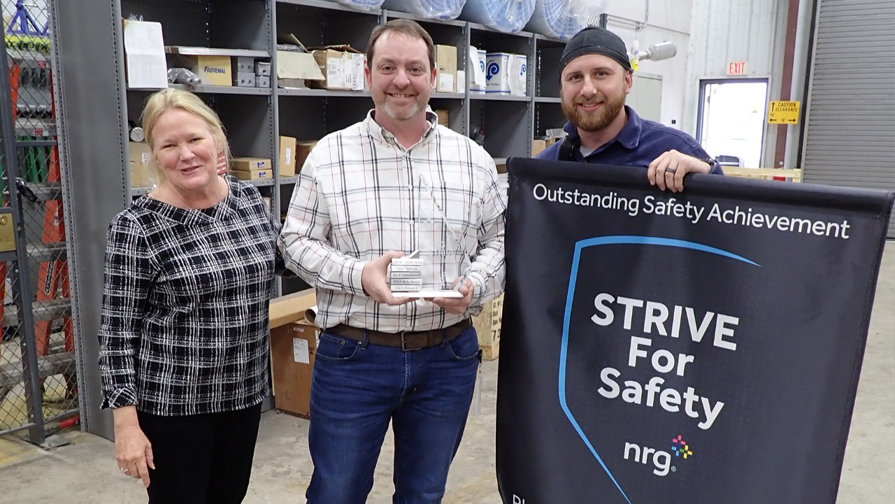 three people stand in a warehouse, one holds a plaque and another holds a Strive for Safety banner.