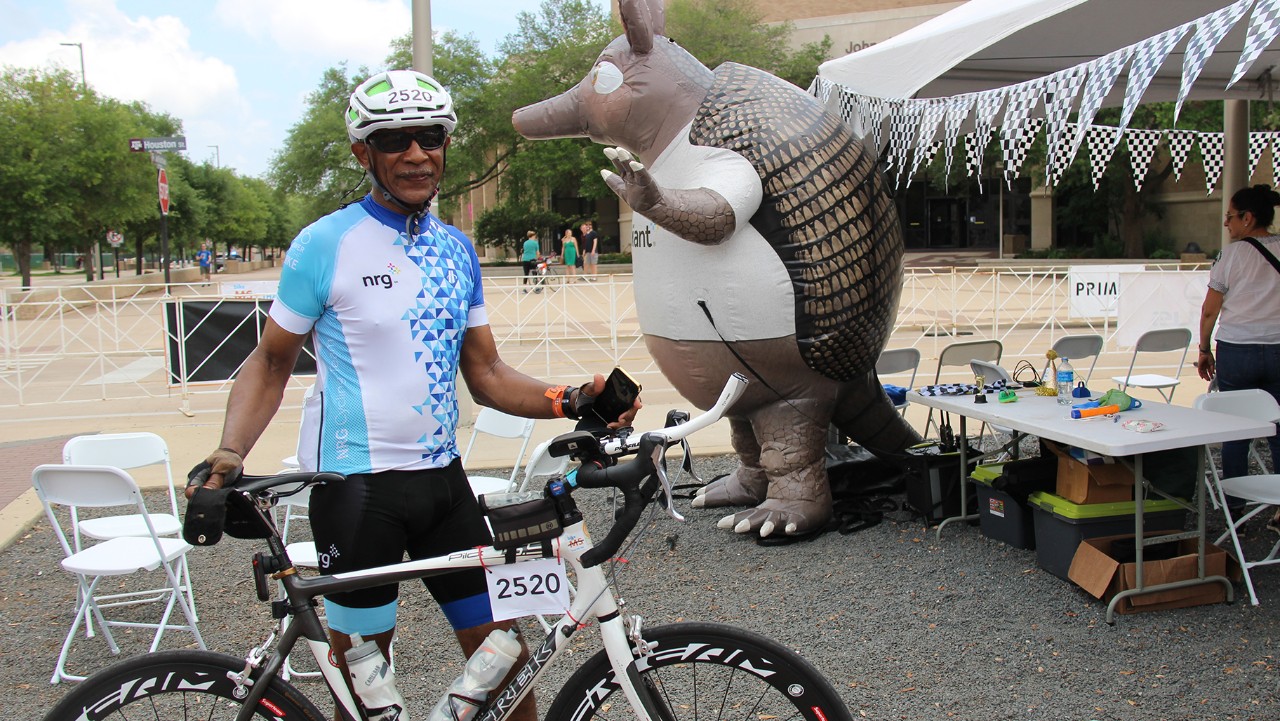 cyclist poses with bike near a large inflatable armadillo at an outdoor event with tents and tables.