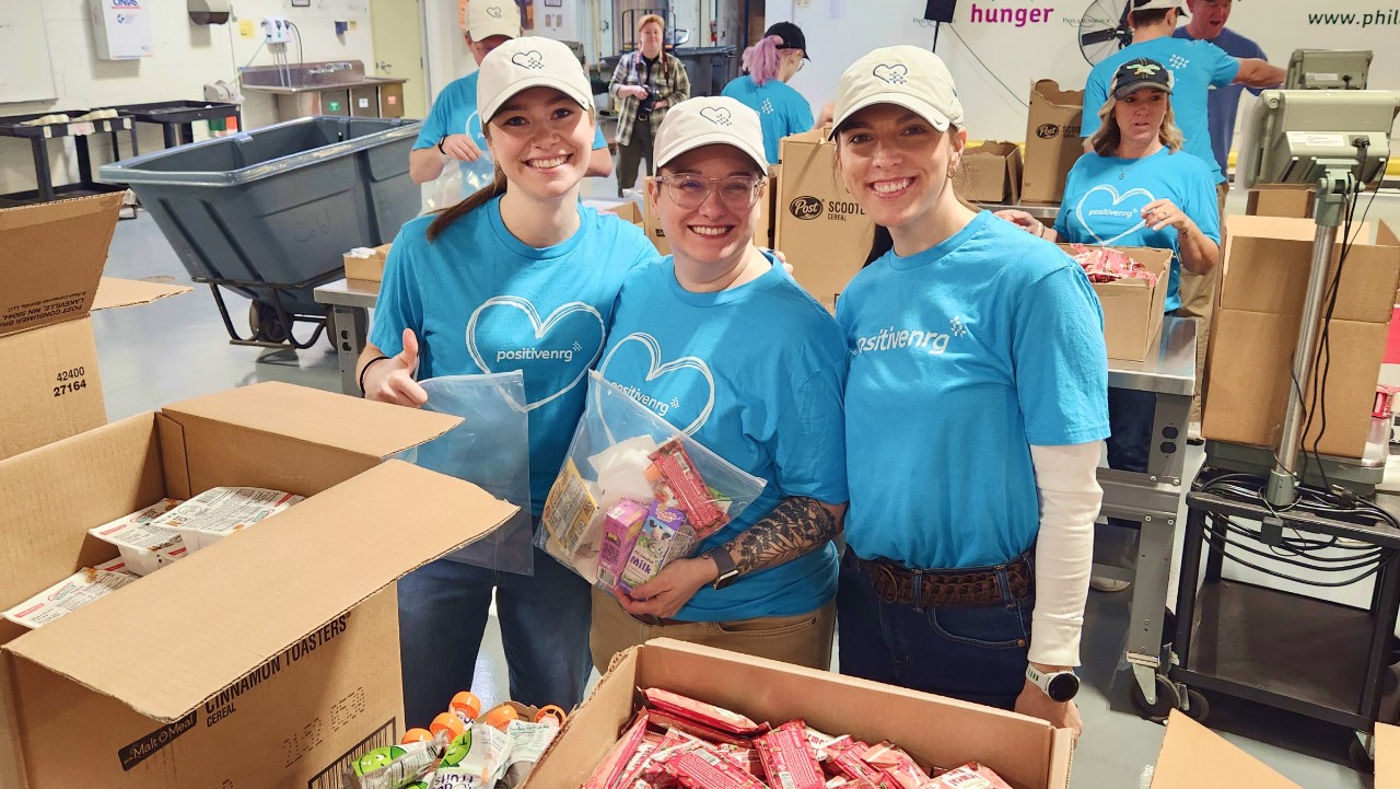 three smiling volunteers pack food items into boxes at a food bank, wearing matching blue shirts and caps.