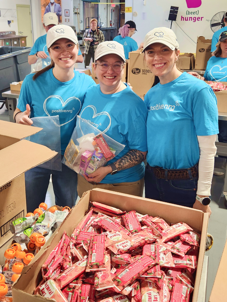 three smiling volunteers in blue shirts pack food items into boxes at a community service event.