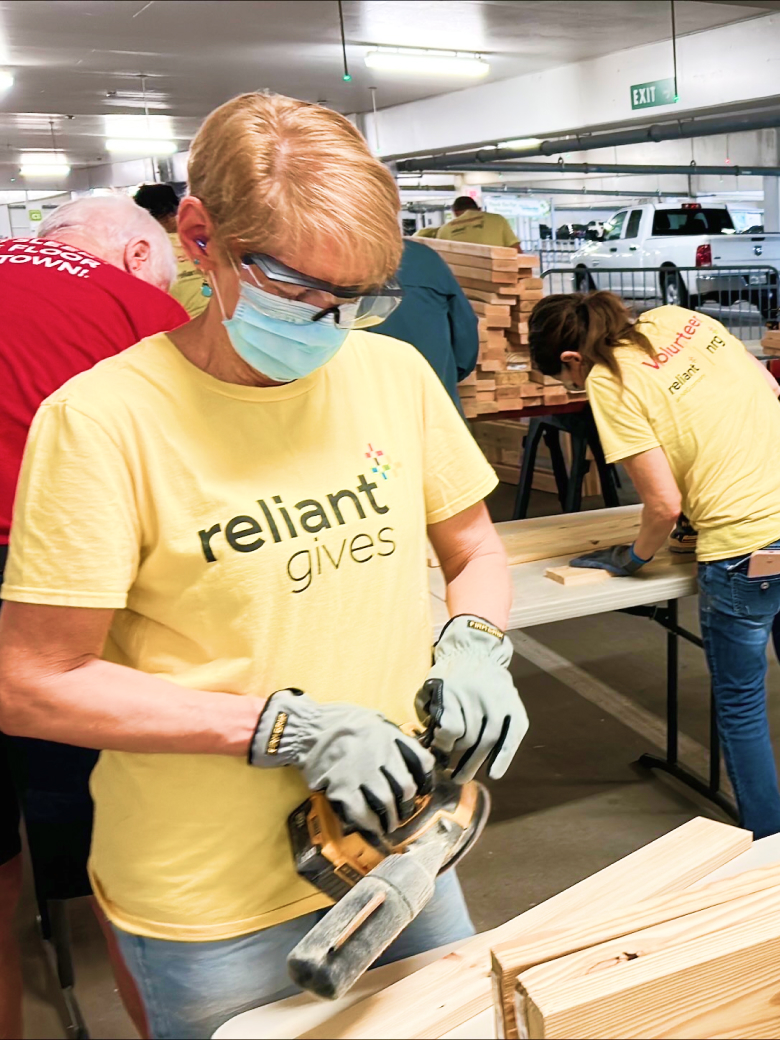 a woman in safety gear sands wood at a table. others wearing reliant gives shirts work nearby.