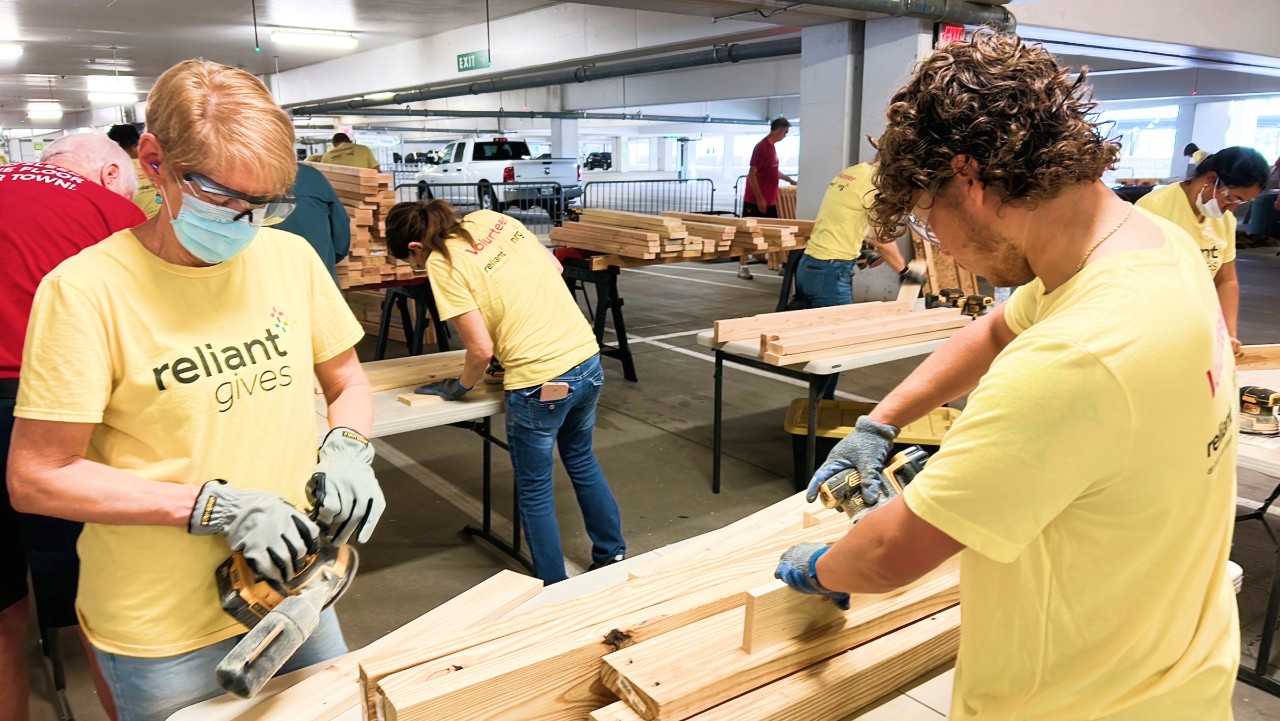 volunteers in yellow shirts use power tools to sand wood planks in a parking garage workspace.