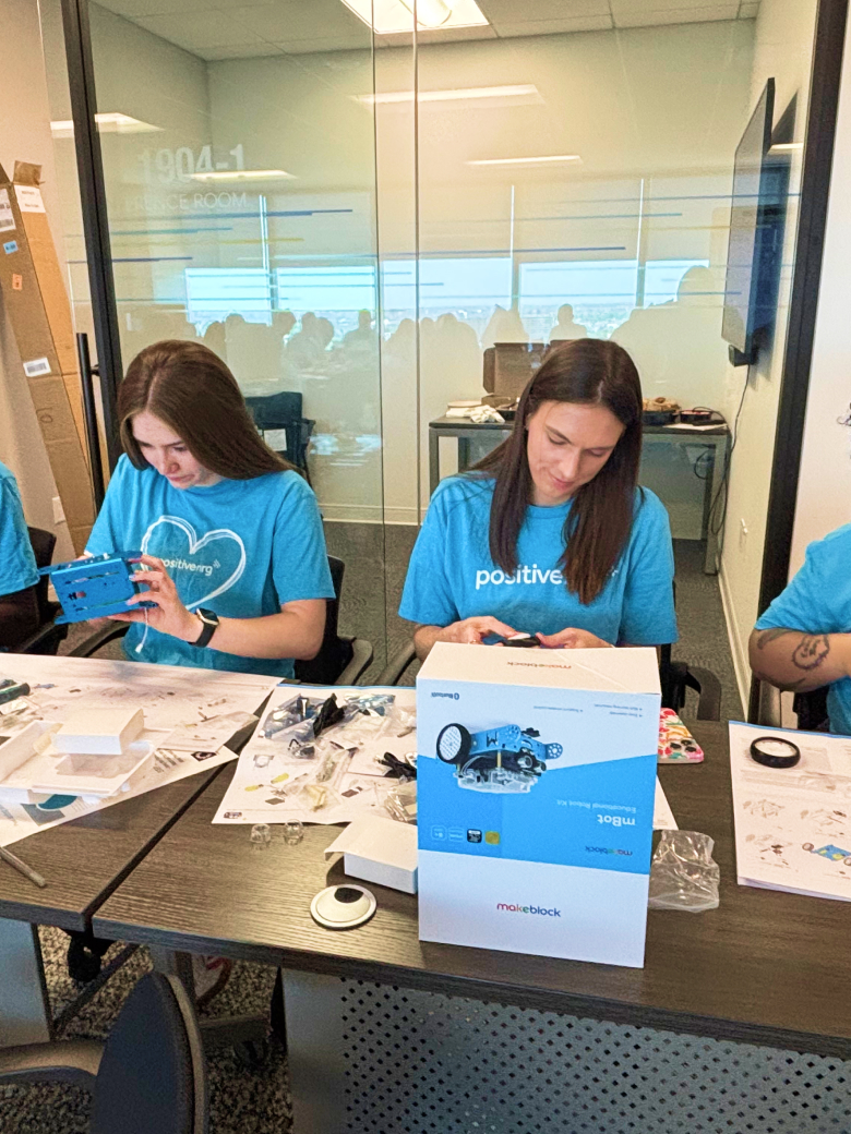 three people in blue shirts assemble robot kits at a table with parts, instructions, and boxes visible.