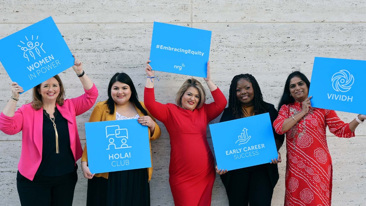 five women stand smiling, each holding a blue sign with different empowering messages and group names.