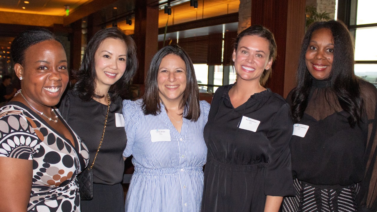 five women standing together indoors, smiling at the camera, each wearing a name tag.