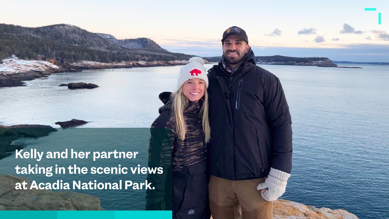 a couple dressed warmly stands smiling on a rocky coast with ocean and hills in the background at sunset.