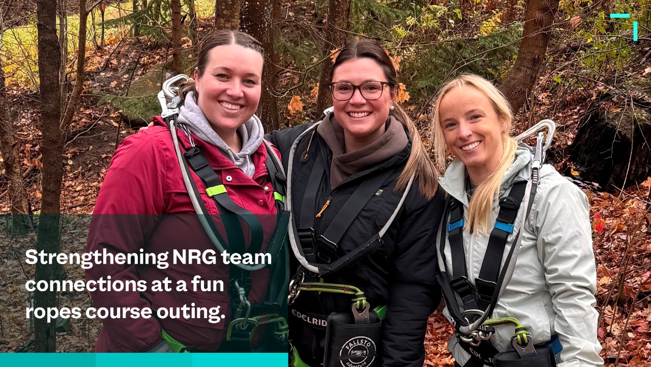 three smiling women in harnesses outdoors at a ropes course, with autumn leaves and trees in the background.