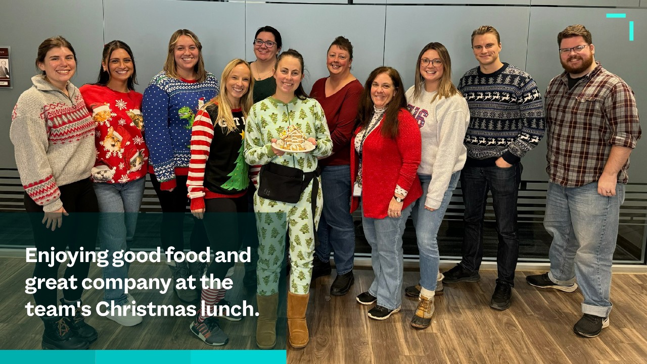 ten people in festive sweaters pose together, smiling, at a Christmas lunch in an office setting.