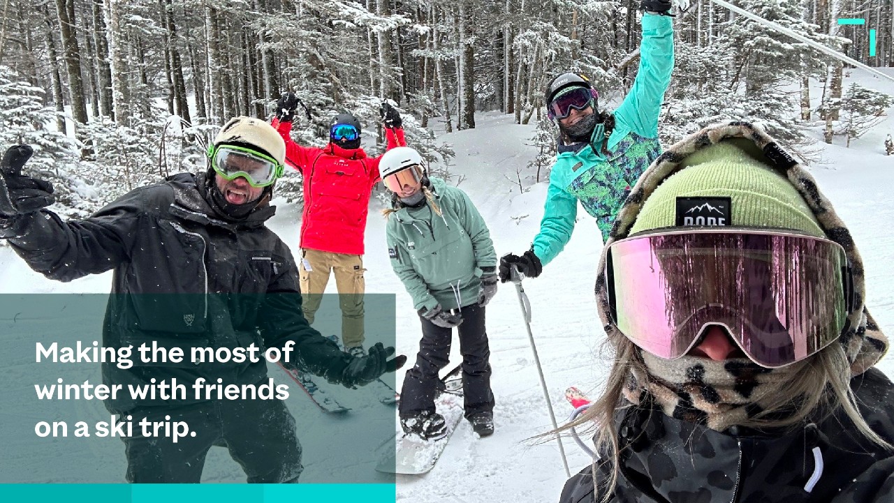 four friends in winter gear pose for a selfie on a snowy ski slope, surrounded by trees.