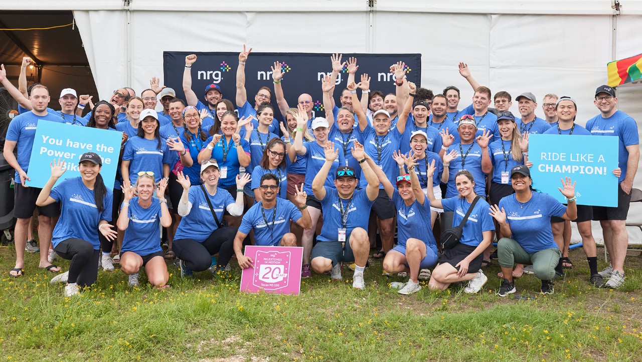 a large group in blue shirts smiles and waves, holding signs, standing in front of a tent with an NRG banner.