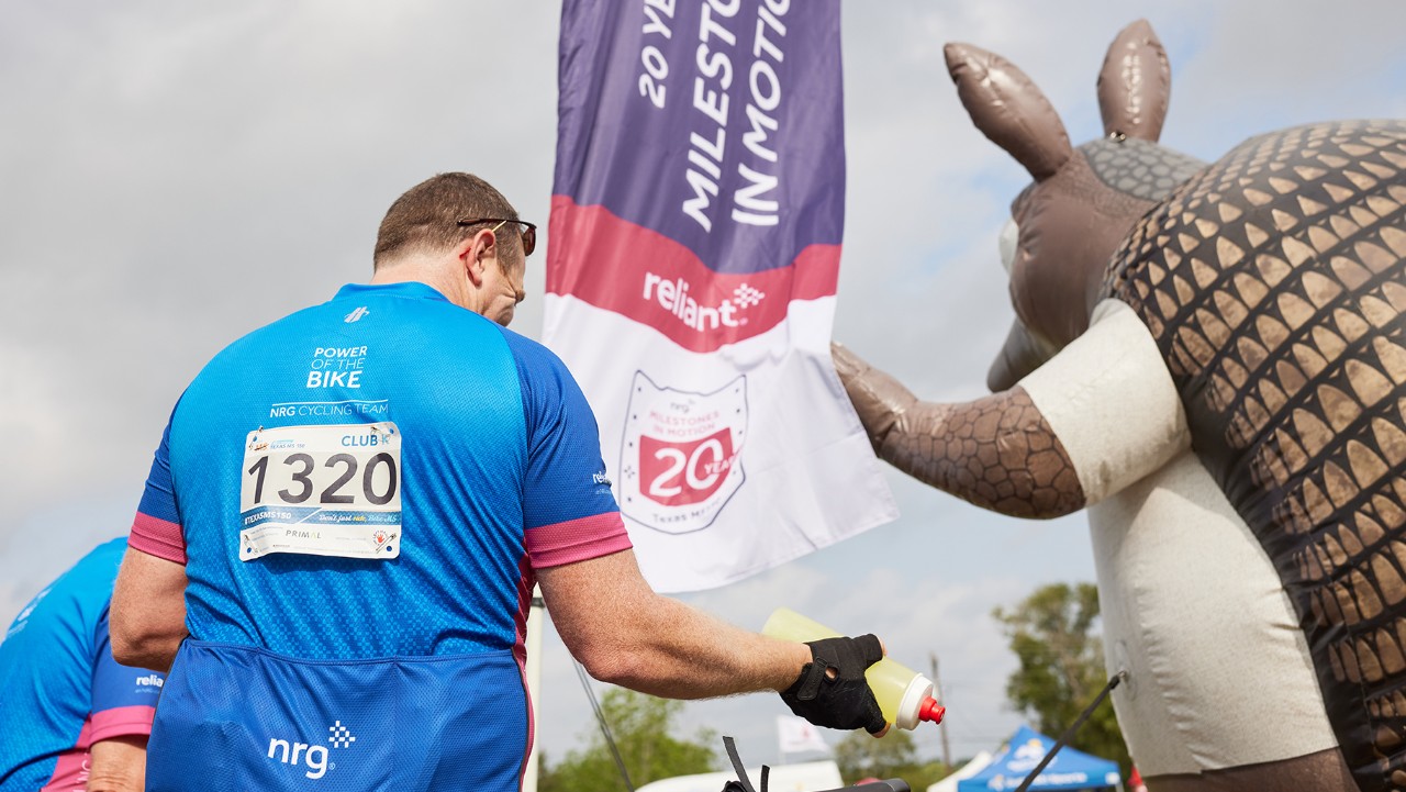 cyclist in blue jersey near large armadillo figure at event, holding a water bottle, event banners visible.