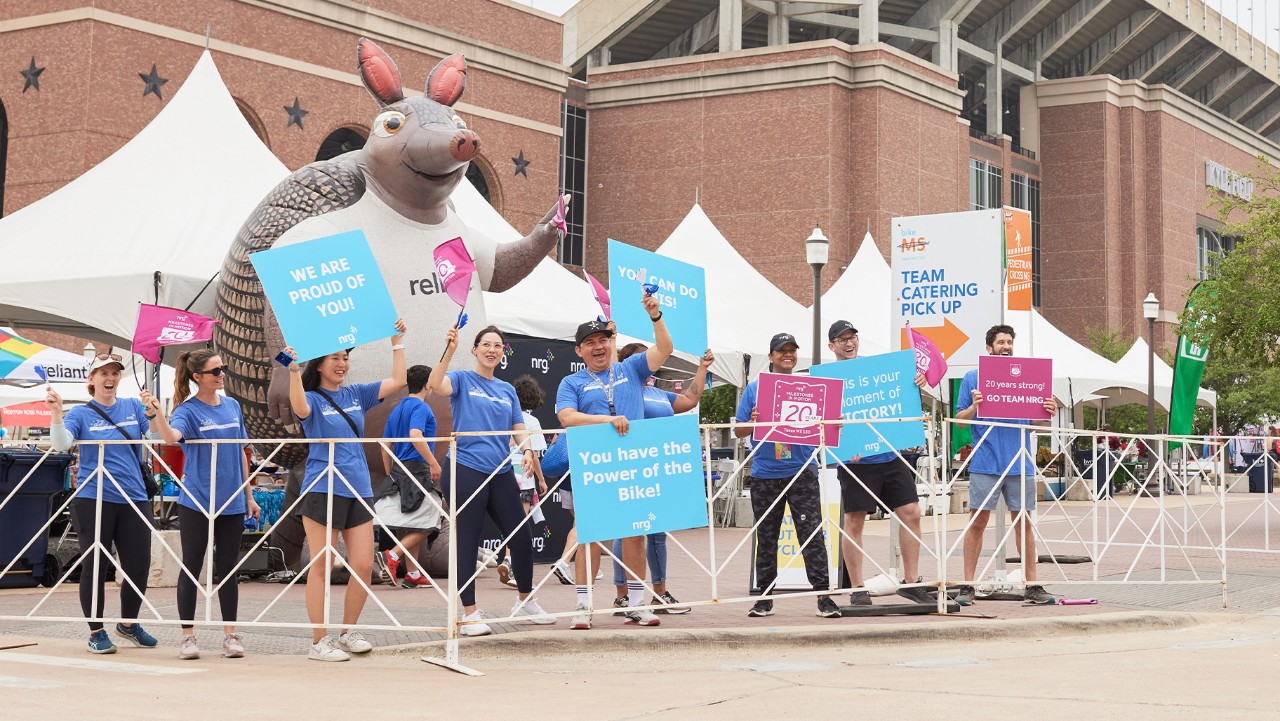 group in blue shirts cheering at an outdoor event with motivational signs and a large armadillo balloon.