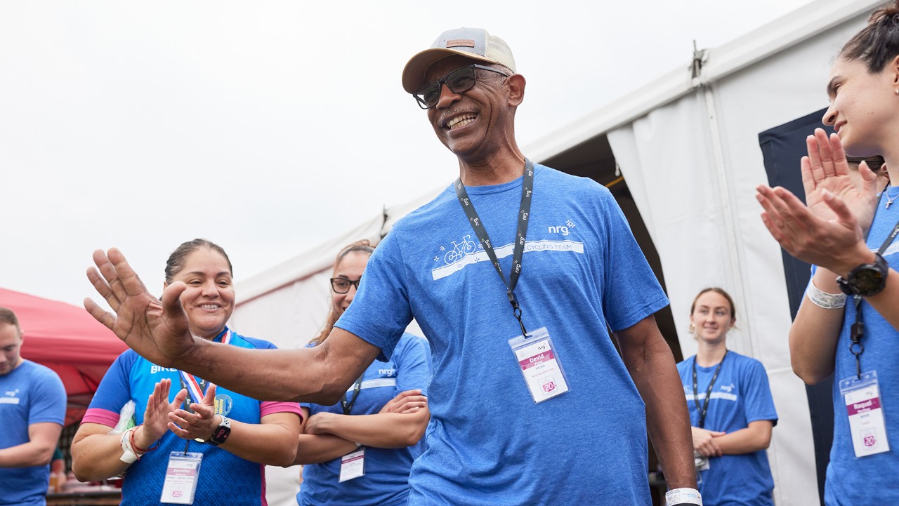 smiling man in blue shirt and cap waves, surrounded by applauding people at an outdoor event.