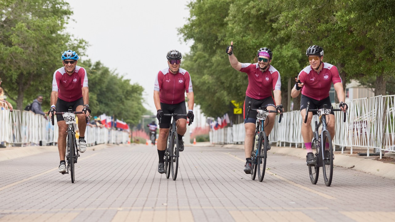 four cyclists in matching maroon jerseys ride together on a tree-lined street during a race event.
