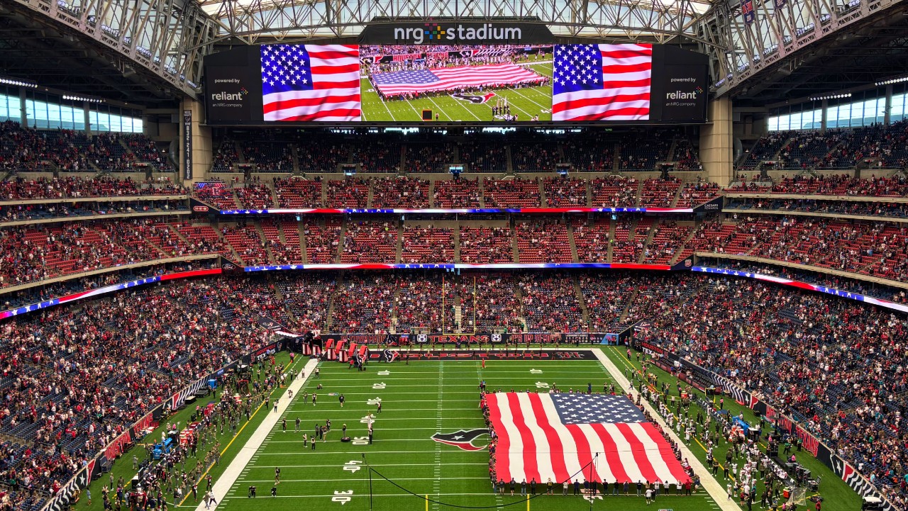large American flag on football field at NRG Stadium, with crowd and scoreboard showing flag display.