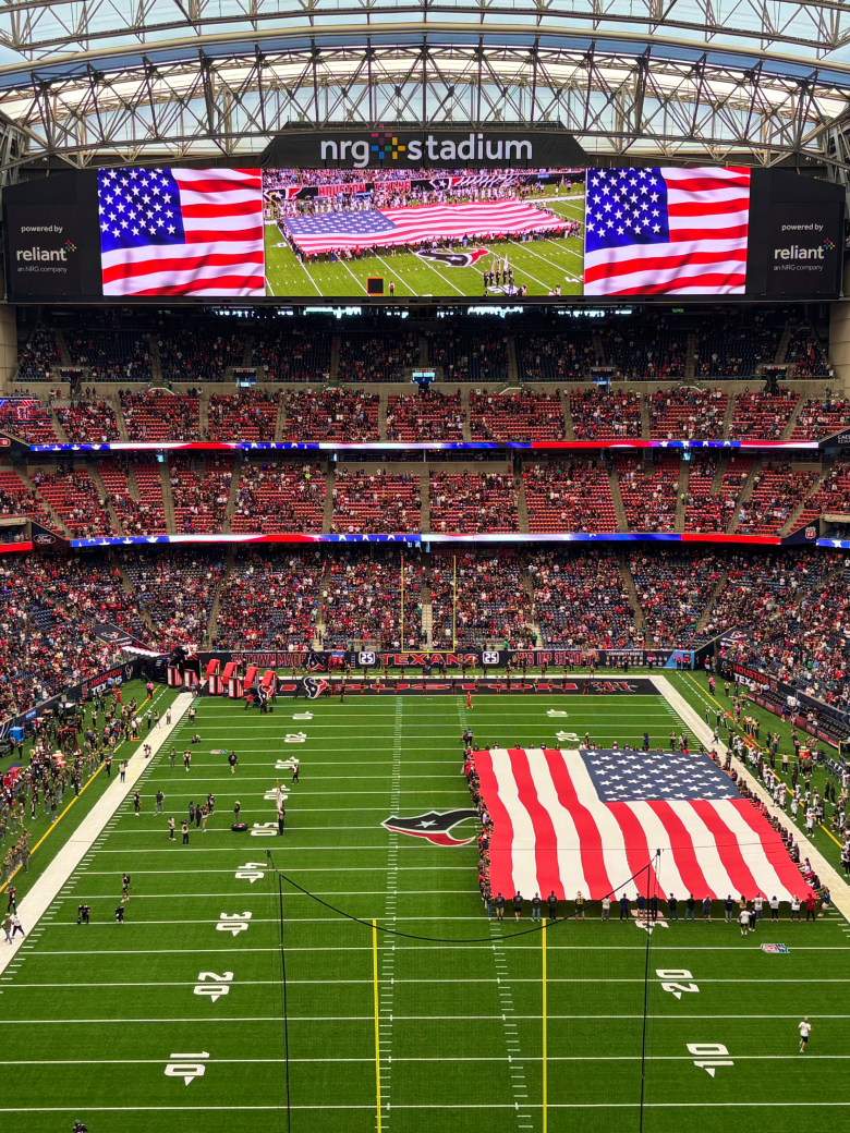 large American flag on football field at NRG Stadium, with fans in stands and flags on the big screen.