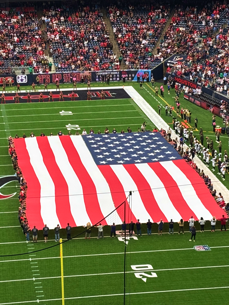 large American flag held on a football field before a game, with crowds in the stadium stands.