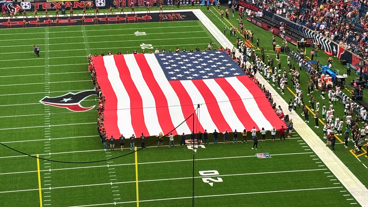 large American flag held by people on a football field before a game, with players lined up on the sidelines.